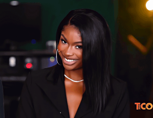 A Woman Wearing A Black Jacket And A Pearl Necklace Smiles In Front Of A Tco Logo GIF