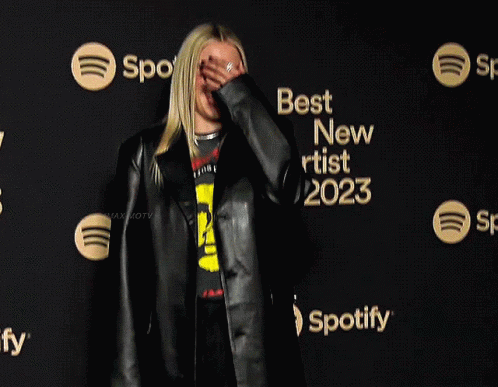 A Woman Wearing A Black Jacket And A Yellow Shirt Stands In Front Of A Sign That Says Best New Artist 2023 GIF