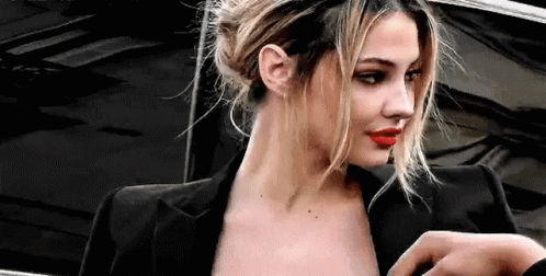 A Woman Wearing A Black Jacket And Red Lipstick Is Standing In Front Of A Black Car . GIF