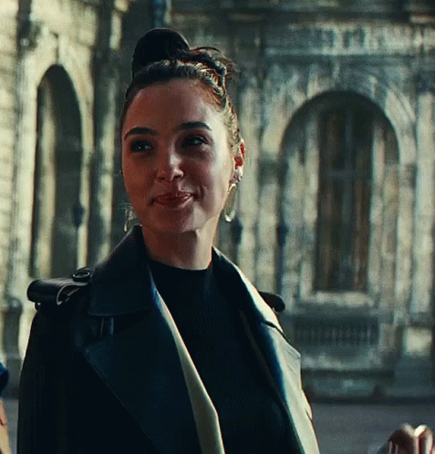 A Woman Wearing A Black Leather Jacket And Earrings Smiles In Front Of A Building GIF