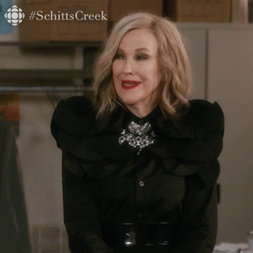 A Woman Wearing A Black Shirt With Ruffles And A Brooch With The Word Schitts Creek On The Bottom Right GIF