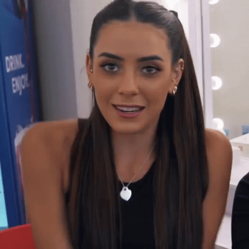 A Woman Wearing A Black Tank Top And A Necklace With A Heart Pendant Is Smiling . GIF