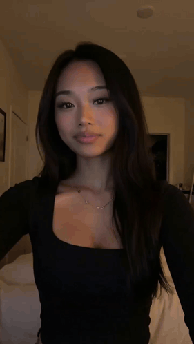 A Woman Wearing A Black Top And A Necklace Is Standing In A Dark Room . GIF