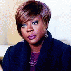 A Woman Wearing A Blue Coat And Earrings Is Sitting On A Couch . GIF