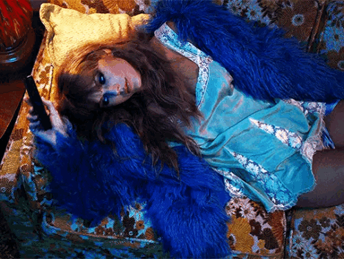 A Woman Wearing A Blue Fur Coat Is Laying On A Couch Holding A Cell Phone . GIF