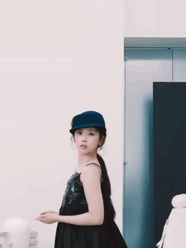 A Woman Wearing A Blue Hat And A Black Dress Looks At The Camera GIF