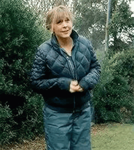 A Woman Wearing A Blue Jacket And Blue Pants Is Standing In Front Of Trees . GIF