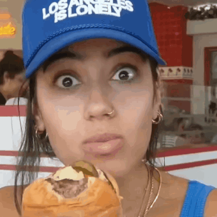 A Woman Wearing A Blue Los Angeles Is Lonely Hat Is Eating A Hamburger . GIF