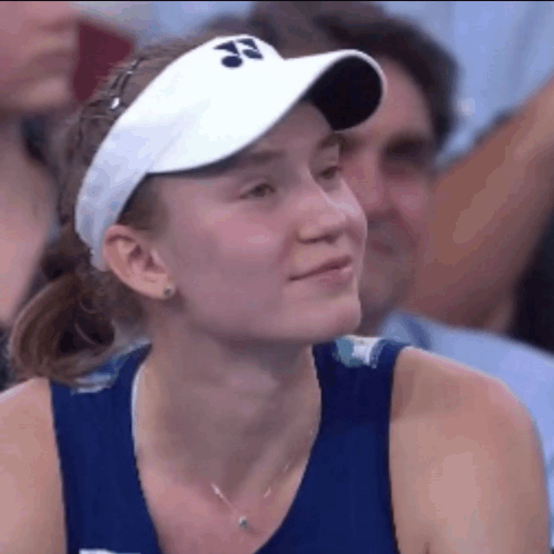 A Woman Wearing A Blue Tank Top And A White Visor Is Smiling While Sitting In A Crowd . GIF