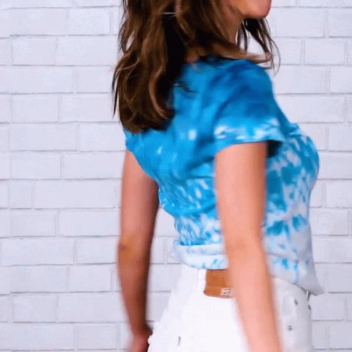 A Woman Wearing A Blue Tie Dye Shirt And White Shorts Is Standing In Front Of A White Brick Wall GIF