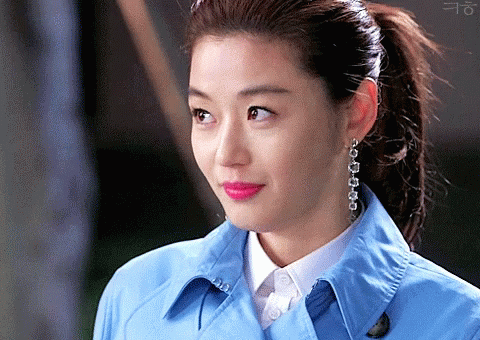 A Woman Wearing A Blue Trench Coat And Earrings Looks At The Camera GIF