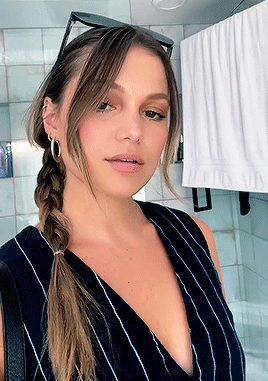 A Woman Wearing A Braided Ponytail And Hoop Earrings Takes A Selfie In A Bathroom GIF