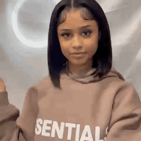 A Woman Wearing A Brown Hoodie Is Standing In Front Of A Wall . GIF