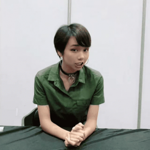 A Woman Wearing A Choker And A Green Shirt Sits At A Table With Her Hands Folded GIF