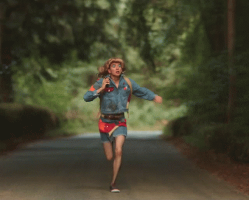 A Woman Wearing A Cowboy Hat And A Denim Jacket Runs Down A Road GIF