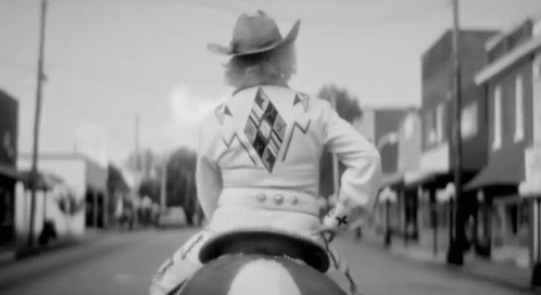 A Woman Wearing A Cowboy Hat Is Riding A Horse Down A Street GIF