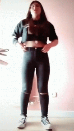 A Woman Wearing A Crop Top And Jeans Is Standing In A Room . GIF