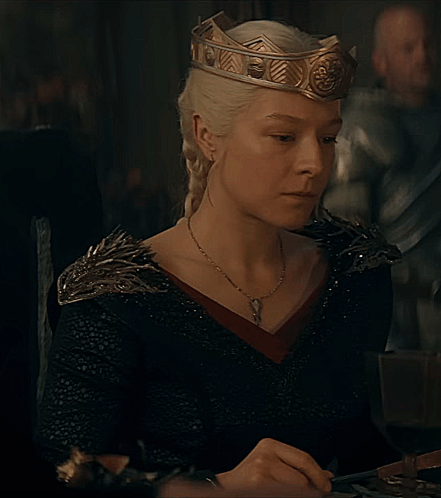 A Woman Wearing A Crown And A Black Dress Is Sitting At A Table GIF
