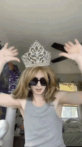 A Woman Wearing A Crown And Sunglasses Holds Her Hands Up In The Air GIF