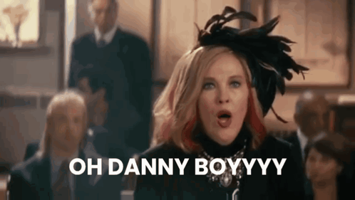 A Woman Wearing A Feathered Hat Is Standing In Front Of A Group Of People And Saying Oh Danny Boyyyy . GIF