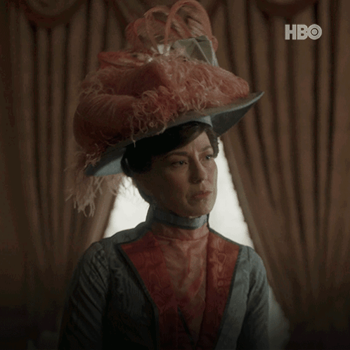 A Woman Wearing A Feathered Hat Says " What Can I Do " In White Letters GIF