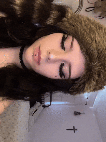A Woman Wearing A Fur Hat And A Choker Has A Cross On The Wall Behind Her GIF
