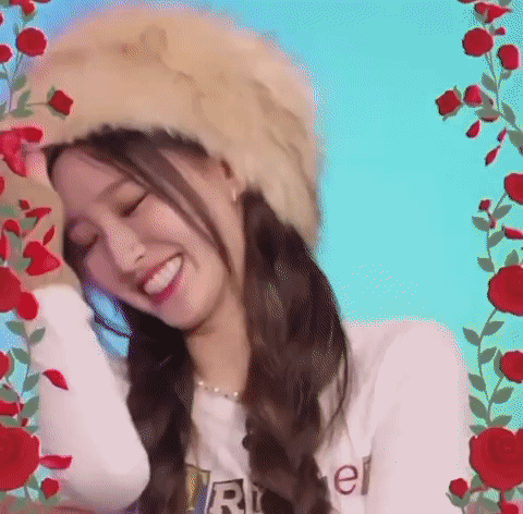 A Woman Wearing A Fur Hat And A White Shirt Is Smiling In Front Of Red Roses . GIF