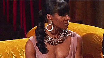 A Woman Wearing A Gold Necklace And Earrings Is Sitting On A Yellow Couch . GIF