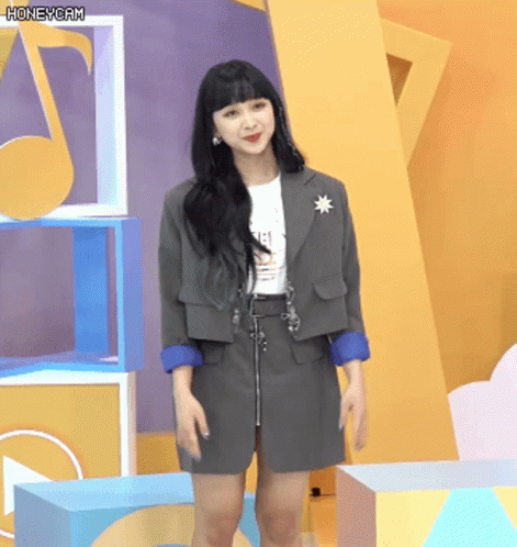 A Woman Wearing A Gray Jacket And Skirt Stands In Front Of A Yellow Wall With A Honeycam Watermark GIF