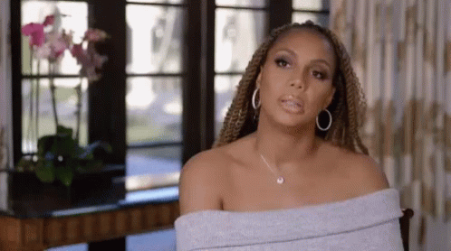 A Woman Wearing A Gray Off The Shoulder Top And Hoop Earrings Is Sitting In A Chair In Front Of A Window . GIF