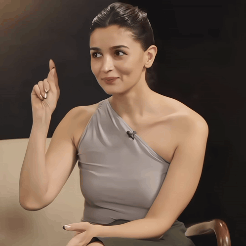 A Woman Wearing A Gray One Shoulder Top Is Pointing Upwards GIF