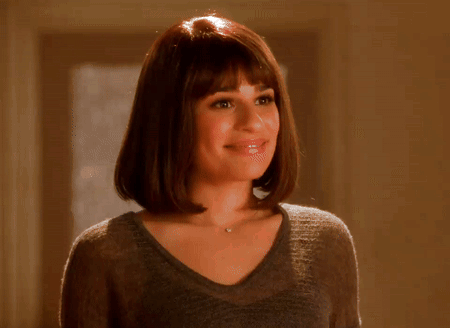 A Woman Wearing A Gray Sweater And A Necklace Smiles GIF