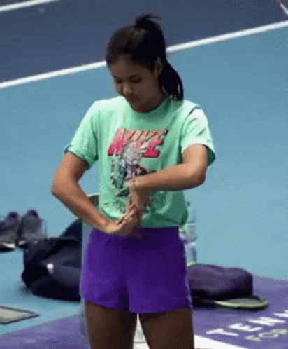 A Woman Wearing A Green Nike T-Shirt And Purple Shorts Is Standing On A Tennis Court . GIF