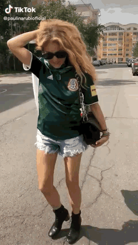 A Woman Wearing A Green Shirt And Shorts Is Standing On The Sidewalk . GIF