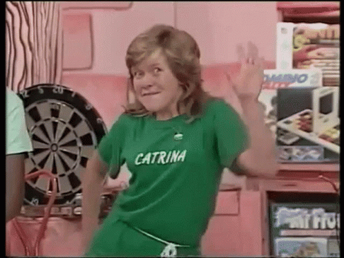 A Woman Wearing A Green Shirt That Says Caterina Is Dancing In Front Of A Dartboard . GIF