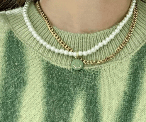 A Woman Wearing A Green Sweater And Pearl Necklaces With A Smiley Face Pendant GIF