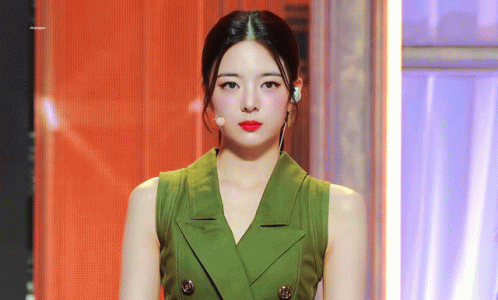 A Woman Wearing A Green Vest And Red Lipstick Is Standing In Front Of A Microphone . GIF