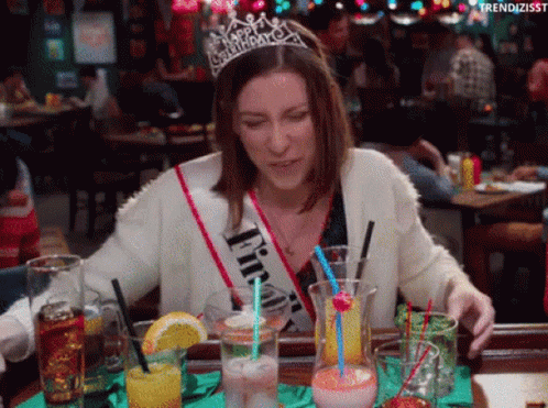 A Woman Wearing A Happy Birthday Sash Is Sitting At A Table With Drinks .. GIF