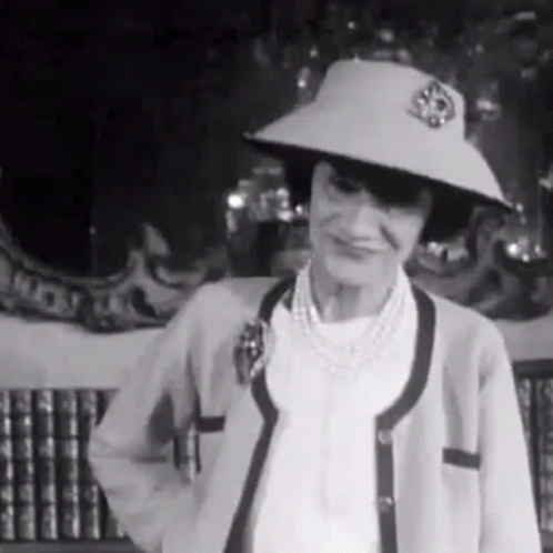 A Woman Wearing A Hat And A Cardigan Is Standing In Front Of A Bookshelf . GIF