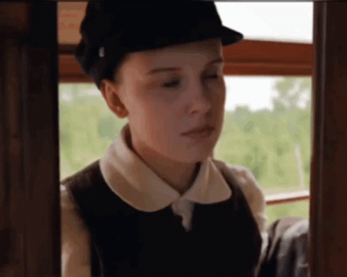 A Woman Wearing A Hat And A Vest Looks Out A Window . GIF