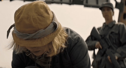 A Woman Wearing A Hat Looks Down At Something While A Man Holding A Gun Stands Behind Her GIF