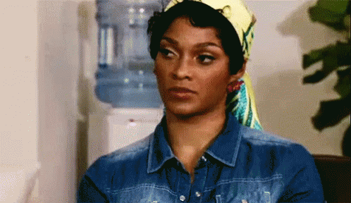 A Woman Wearing A Head Scarf And A Denim Shirt Is Standing In Front Of A Water Cooler . GIF