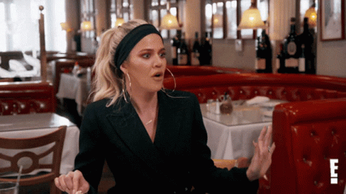 A Woman Wearing A Headband And Hoop Earrings Is Sitting At A Table In A Restaurant With The Letter E In The Corner GIF