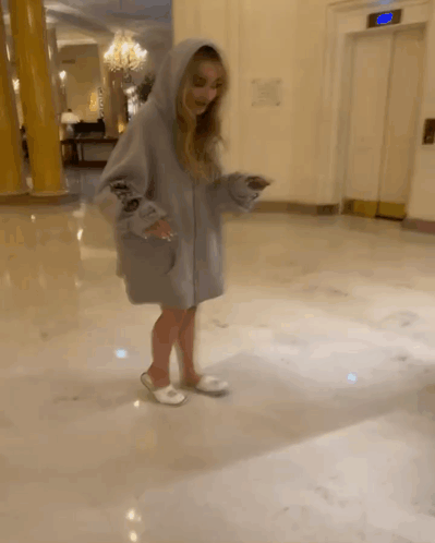 A Woman Wearing A Hoodie And Slippers Is Standing In A Hallway Next To An Elevator GIF