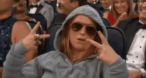 A Woman Wearing A Hoodie And Sunglasses Is Sitting In A Crowd Of People And Making A Peace Sign . GIF