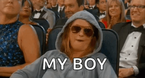 A Woman Wearing A Hoodie And Sunglasses Is Sitting In A Crowd Of People And Says `` My Boy '' . GIF