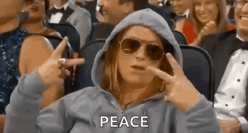 a woman wearing a hoodie and sunglasses is sitting in a crowd of people making a peace sign .