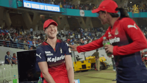 A Woman Wearing A Kalaria Shirt Talks To Another Woman In A Red Hat GIF