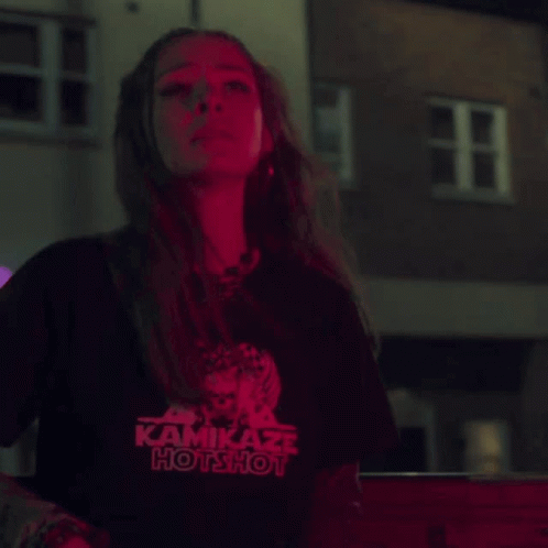 A Woman Wearing A Kamikaze Hotshot T-Shirt Stands In Front Of A Building GIF