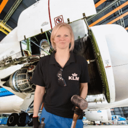 A Woman Wearing A Klm Shirt Holds A Hammer In Front Of An Airplane GIF